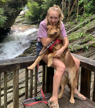 Lily Sronkoski holding her dog with a forest and stream in the background