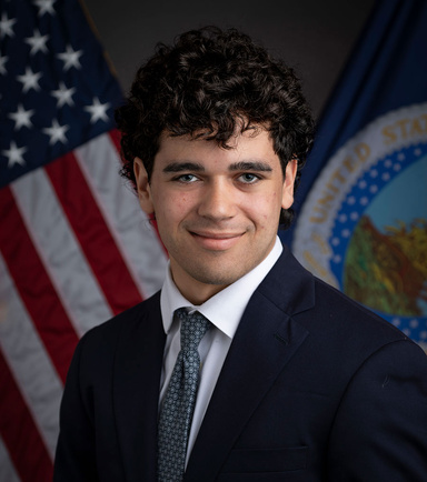 Photo of Joe Maxwell, wearing a dark suit and the American flag behind him. 