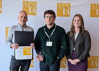 Three individuals accepting the award infront of a background with yellow APA logos.