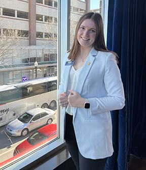 Airiana Mohr, wearing a light blue suit, standing by a window showing a city view with a tall building plus bus and car traffic below.