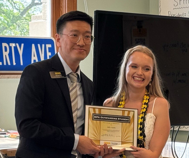 Allie Tubbs, wearing a white lacey dress, standing next to Haifeng Qian as she receives the framed award.