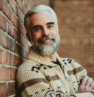 Kyle M. Smith wearing a sweater and leaning against a brick wall. 