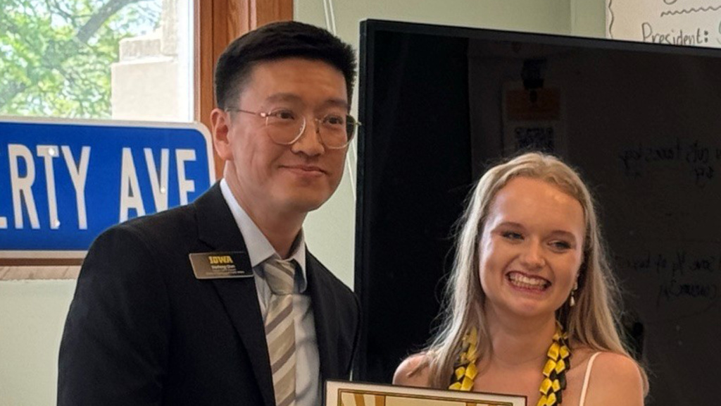 Allie Tubbs, wearing a white lacey dress, standing next to Haifeng Qian as she receives the framed award.